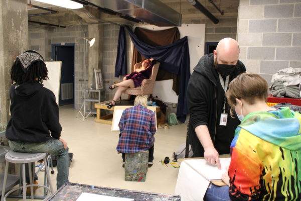 Instructor Evan Kitson works with students in a life model class; model seated out of focus in background.