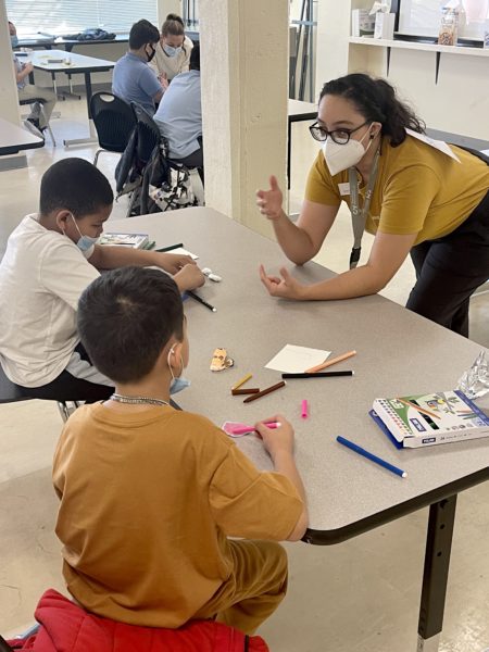 woman with long dark hair works with two 5th-graders who are coloring clay models.