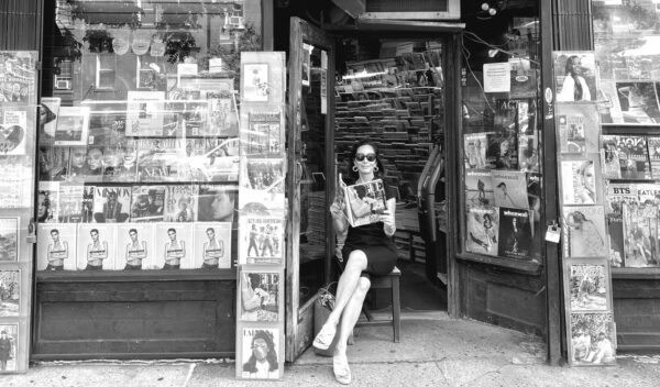 black and white of classically dressed woman in sunglasses sitting in front of NYC shop.
