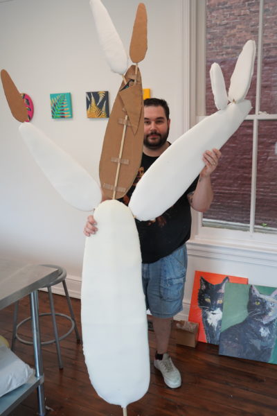 bearded man in black shirt stands behind the cardboard and wire frame of a giant cactus sculpture.