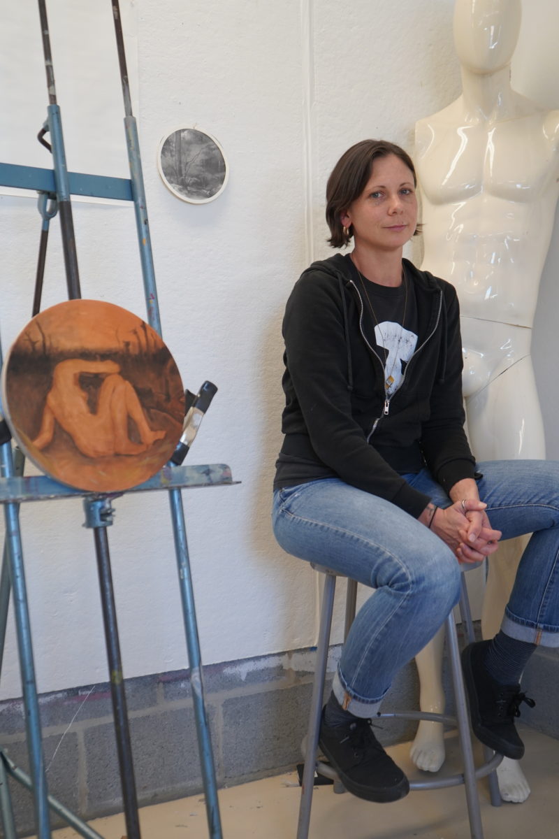 woman with short brown hair sits on stool and looks off to the right, smiling slightly at camera, with her oval art in progress on easel to her right. 