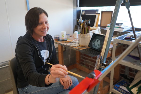 woman with short brown hair smiling and holding paintbrush as she works on oval canvas.