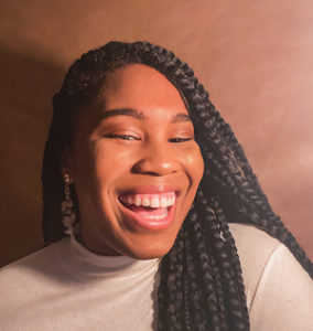 Young Black woman with braids over her left shoulder smiling at camera against brown background.