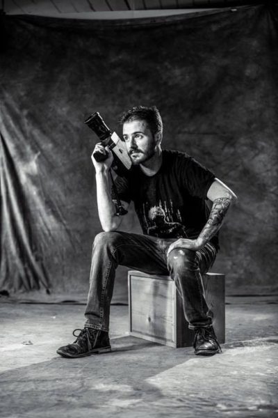 black and white photo of man sitting on box holding camera above right shoulder. 