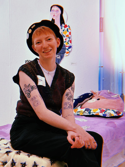 three-quarter shot of artist with red hair wearing beret seated in front of a portrait by the artist on a white wall.