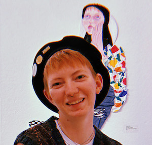 headshot of person with red hair wearing a beret, with a portrait by the artist behind them on a white wall.