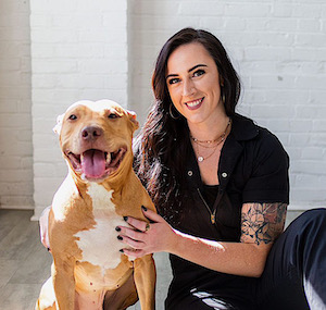 Woman with long dark hair in black short sleeve shirt posing with smiling tan dog