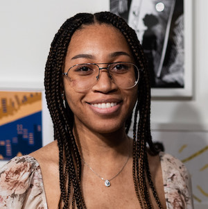 Woman with long dark braids and eyeglasses smiling in front of her artwork.