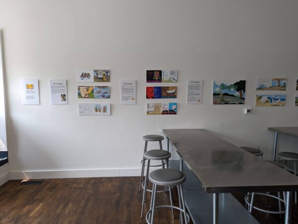 Groupings of artwork on a white wall with a metal table and stools in front.