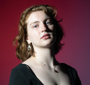 headshot dramatically lit of woman in scoopneck black top against maroon background.