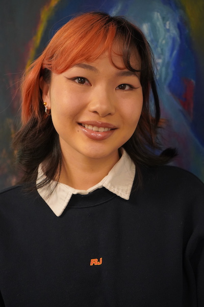 Young woman with red and black hair, dark sweater and white blouse smiling for camera.